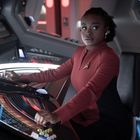 Eine Frau in rotem Uniform sitzt vor einem futuristischen Bedienfeld, umgeben von Technologie in einem Raumschiff.