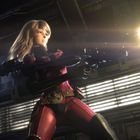 Eine Frau in rotem Anzug hält eine große Waffe. Sie steht in einer metallischen, düsteren Umgebung.