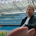 Ein Mann in schwarzer Jacke lächelt in einem leeren Stadion. Im Hintergrund sichtbar: Sitzreihen und ein großes Dach.