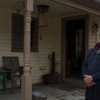 Eine Person steht auf einer überdachten Veranda, trägt eine dunkle Jacke und eine Brille. Daneben ein Tisch und ein Stuhl.