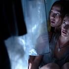 Zwei junge Frauen kauern ängstlich in einer Ecke, von schwachem, blauem Licht beleuchtet. Plastikhülle im Vordergrund.