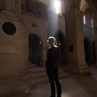 Ein Mensch steht in einer alten Steinhalle mit hohen Rundbogenfenstern, durch die Sonnenstrahlen fallen.