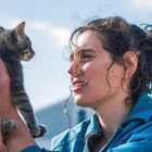 Eine Person hält ein graues Kätzchen in den Händen, lächelnd und in blauem Oberteil, vor einem hellen, wolkigen Himmel.