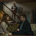 Ein Mann und eine Frau sitzen auf einem Sofa vor einem Tisch mit Gläsern. Eine weitere Person steht auf der Treppe.