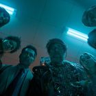 Sechs Menschen schauen ausdruckslos nach unten, beleuchtet von blauem Licht. Einer trägt einen Drahtanzug.