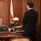 Zwei Männer in einem eleganten Büro, einer sitzt hinter einem Schreibtisch mit russischer Flagge im Hintergrund.