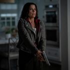 Keine sichtbaren Texte. Eine Frau mit dunklem Haar hält eine Pistole in einer Küche, wirkt besorgt. Sie trägt eine Jacke.