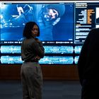 Text: "James Bonomo." Ein Raum mit großen Bildschirmen, eine Frau steht nachdenklich davor.