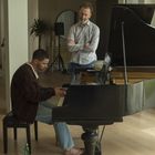 Ein Mann spielt Klavier, während ein anderer daneben steht. Im Hintergrund sind weitere Personen und ein Fenster zu sehen.