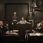 Eine ernste Gerichtsszene mit Uniformierten, Personen tragen Kopfhörer, ein Mann steht im Fokus vor einer Tür mit Wappen.