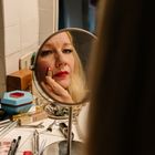 Eine Frau sieht in einen Spiegel, während sie roten Lippenstift aufträgt. Um sie herum liegen viele Schminkutensilien.