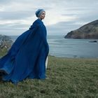 Eine Person in einem blauen Umhang steht auf einer Wiese vor einer Küstenlandschaft mit Meer und Hügeln.