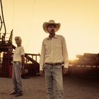 Zwei Männer stehen auf einer Baustelle bei Sonnenuntergang. Der eine trägt einen Cowboyhut, der andere ein weißes T-Shirt.