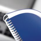 Blaues Spiral-Notizbuch mit weißer Spirale und sichtbarer Kante. Hintergrund unscharf, vermutlich eine Tastatur.