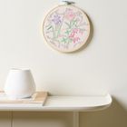 Eine runde Stickerei zeigt bunte Blumen an der Wand über einem schwebenden Regal mit weißer Vase und Buch.