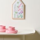 Stickerei von Blumen in einem holzgerahmten Bild an der Wand über zwei rosa Kerzenhaltern auf weißem Regal.