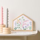 Eine Holzplatte in Hausform mit gestickten Blumen, daneben spiralförmige Kerzen in Rosa und Grau auf Holzständern.