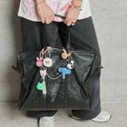 Text: "Balenciaga". Große schwarze Tasche mit gestrickten Tier-Anhängern, getragen von einer Person in lässiger Kleidung.