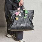 Großes schwarze Tasche mit gehäkeltem Blumenschmuck, gehalten von Hand. Träger trägt dunkle Hose und helle Turnschuhe.