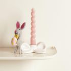Ein filzgrauer Hase hält ein buntes Ei neben einer gedrehten rosa Kerze auf einer weißen, schleifenförmigen Halterung.