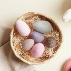 Ein Korb mit bunten, pastellfarbenen Eiern auf Holzwolle, daneben weiße Tulpen und ein rosa Teller.