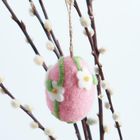 Ein rosa Filzei mit weißen Blüten hängt an einem Ast mit flauschigen Knospen.