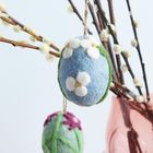 Zwei Filzeier mit Blumenmuster hängen an Weidenzweigen in einer rosafarbenen Vase.