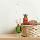 Kleiner Plüschhase in grün mit Karotte neben einem Korb voller Filzkarotten.