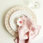 Ein weißer Teller auf einem geflochtenen Untersetzer, darauf eine rosa Serviette mit einer blumenförmigen Spange. Ein Glas Wasser.