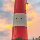 Ein rot-weißer Leuchtturm steht majestätisch vor einem bewölkten Himmel, umgeben von Dünen und Pflanzen. Unten ist ein Kalender.