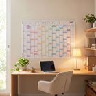 Ein Wandkalender für 2027 hängt über einem Schreibtisch mit Laptop, Pflanze und Lampe, daneben sind Bücherregale.