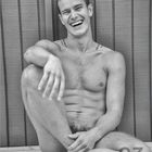 März. Ein lächelnder, nackter Mann sitzt entspannt an eine Holzverkleidung gelehnt.