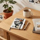 Auf einem Holztisch liegt ein Notizbuch mit "Einfach mit dem Mond". Daneben eine Tasse Tee, ein Stift, Bücher und eine Pflanze.