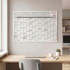 Ein Wandkalender für 2027 hängt über einem Holztisch mit Laptop, Tasse und Pflanze. Regale sind im Hintergrund.