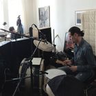 Ein Musiker spielt Schlagzeug, umgeben von Tonstudio-Ausstattung. Im Hintergrund ist eine Hexenfigur zu sehen.