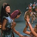 Drei Frauen in eleganter Kleidung; eine im Hintergrund hält einen Spiegel, der auf eine Vase mit Blumen gerichtet ist.