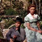 Ein Mann mit Hut sitzt neben einer Frau in einem grünen Kleid vor einem Hintergrund aus Pflanzen und Felsen.