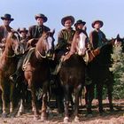 Sieben Cowboys auf Pferden, alle tragen Hüte, dicht nebeneinander auf staubigem Boden.