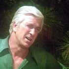 Ein Mann mit grauem Haar in einem grünen Hemd steht vor einem Hintergrund aus Tannenzweigen.