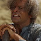 Eine Person mit zerzaustem Haar hält einen schmutzigen Stock. Der Hintergrund wirkt sandig und trocken.