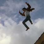 Ein Mann springt mit ausgestreckten Armen von einem Gebäude, hält eine Pistole, vor bewölktem Himmel.