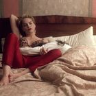Eine Frau in rotem Outfit und Leopardenmuster-Oberteil sitzt lässig im Bett, Arm hinter dem Kopf, weiche Beleuchtung.
