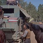 Ein Mann sitzt auf einem Pferdewagen, redet mit einem Reiter und einem Jungen. Im Hintergrund Wald und klarer Himmel.