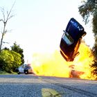 Ein Auto in der Luft, daneben eine Explosion. Ein Polizeiauto befindet sich auf der Straße, umgeben von Bäumen.