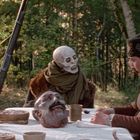 Ich sehe keinen Text. Ein Skelett und eine Person sitzen an einem Tisch im Wald. Auf dem Tisch liegt ein künstlicher Kopf.