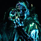 Drei Personen mit fluoreszierender, bunter Bemalung auf dunklem Hintergrund. Gesichter und Kleidung leuchten intensiv.