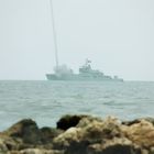 Kein Text erkennbar. Ein Kriegsschiff auf dem Meer feuert eine Rakete ab, im Vordergrund Felsen im Wasser.