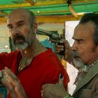 Zwei Männer, einer bedroht den anderen mit einer Pistole am Kopf. Der Bedrohte trägt Rot und wirkt angespannt.