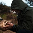 Ein Mann in olivgrünem Parka betrachtet einen Schädel mit einem Metallkreuz darin in einer bewaldeten Umgebung.