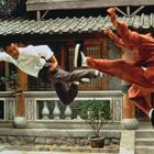 Zwei Menschen kämpfen in der Luft, einer in weiß und der andere in rot, vor traditioneller asiatischer Architektur.
